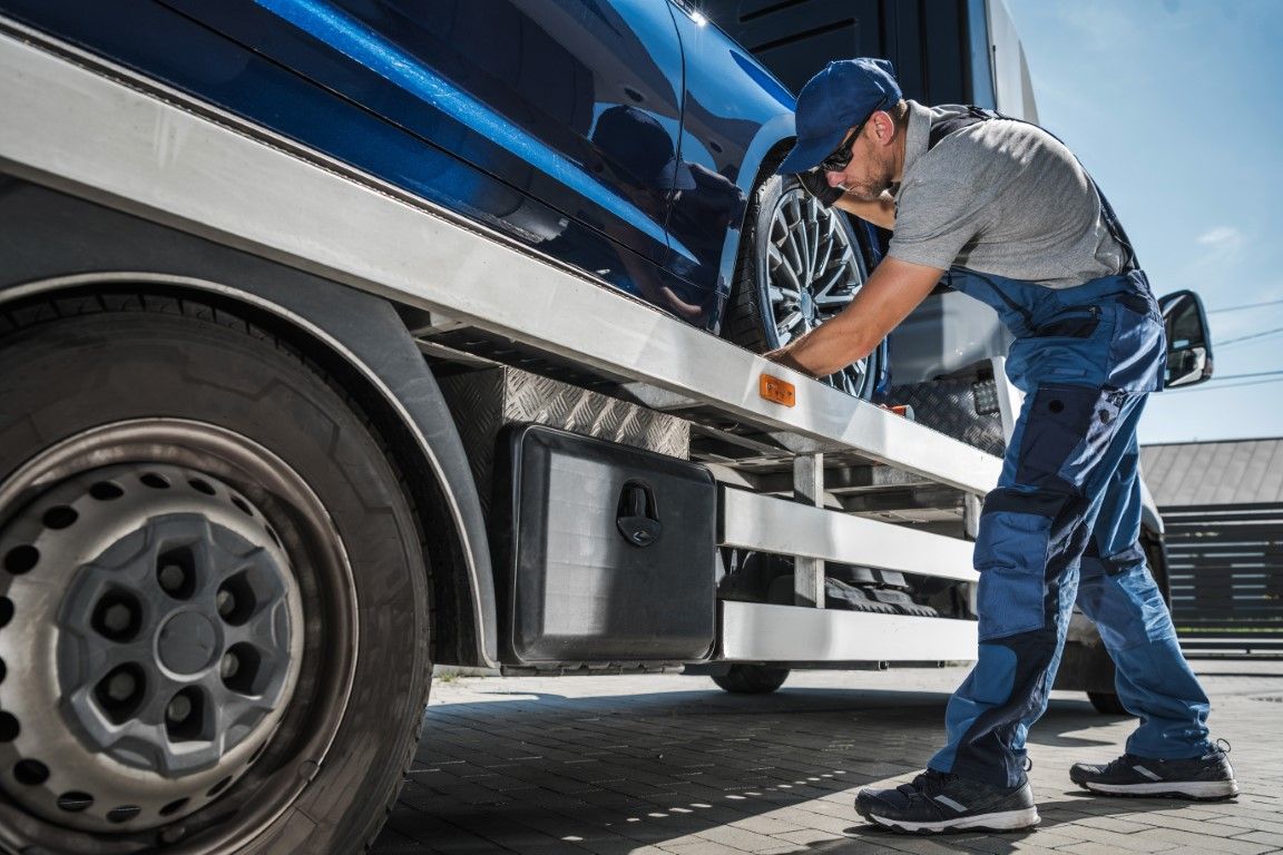 Tow truck operator secures a blue van onto the flatbed. Outdoors, sunny.