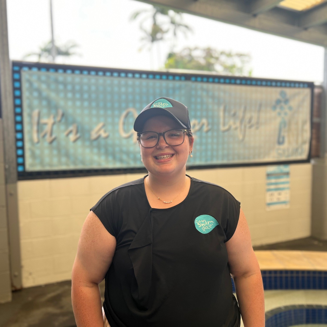 Smiling person in black cap and shirt stands indoors beside a tiled pool and decorative wall sign.