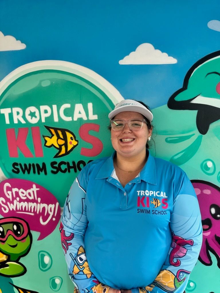 Smiling person in black cap and shirt stands indoors beside a tiled pool and decorative wall sign.