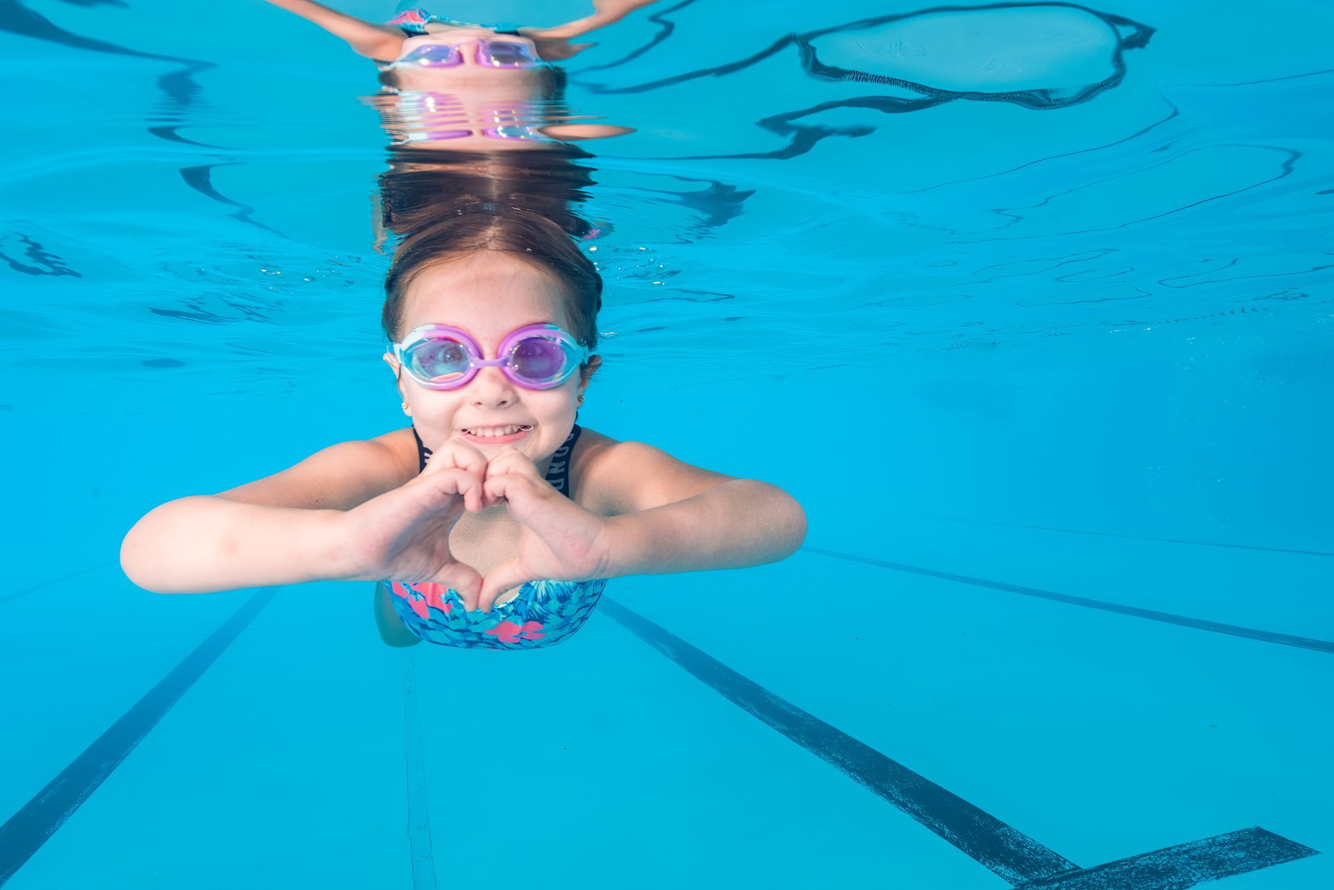 Child swimming underwater in a pool, wearing pink goggles and a striped swimsuit, smiling at the camera