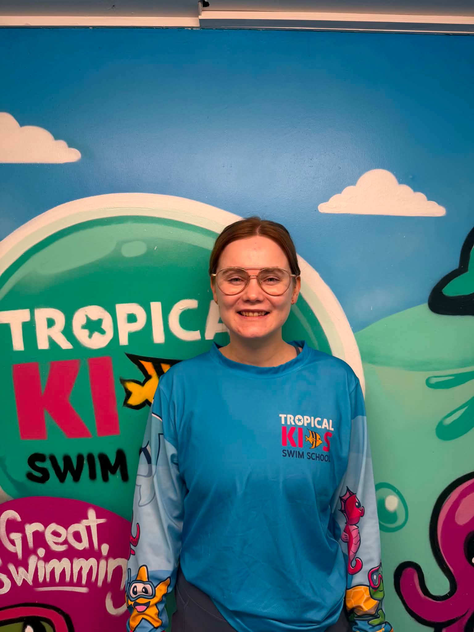 Smiling person in black cap and shirt stands indoors beside a tiled pool and decorative wall sign.