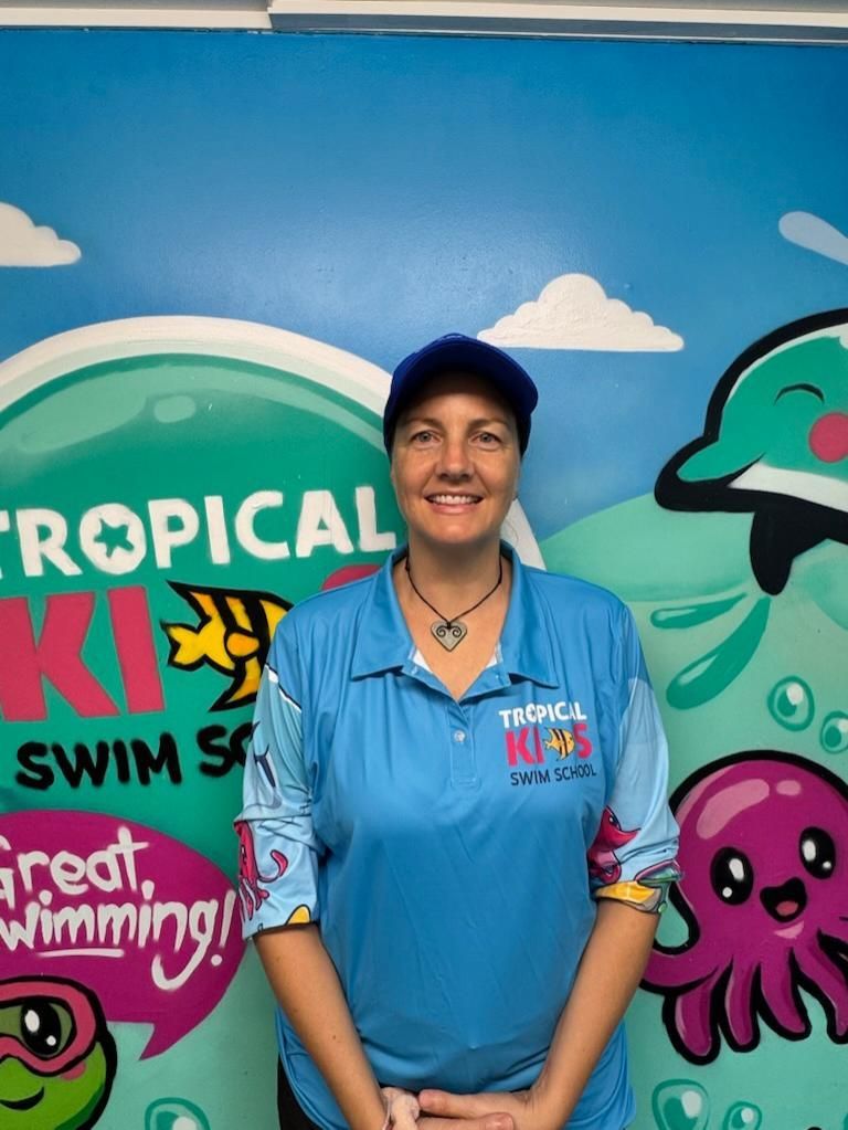 Smiling person in black cap and shirt stands indoors beside a tiled pool and decorative wall sign.