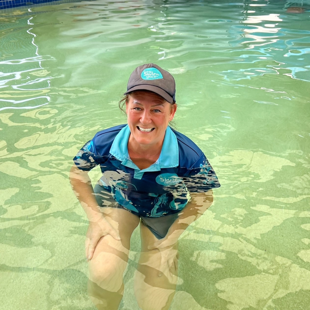 Person smiling in a pool, wearing a cap and blue shirt, standing in clear green water.