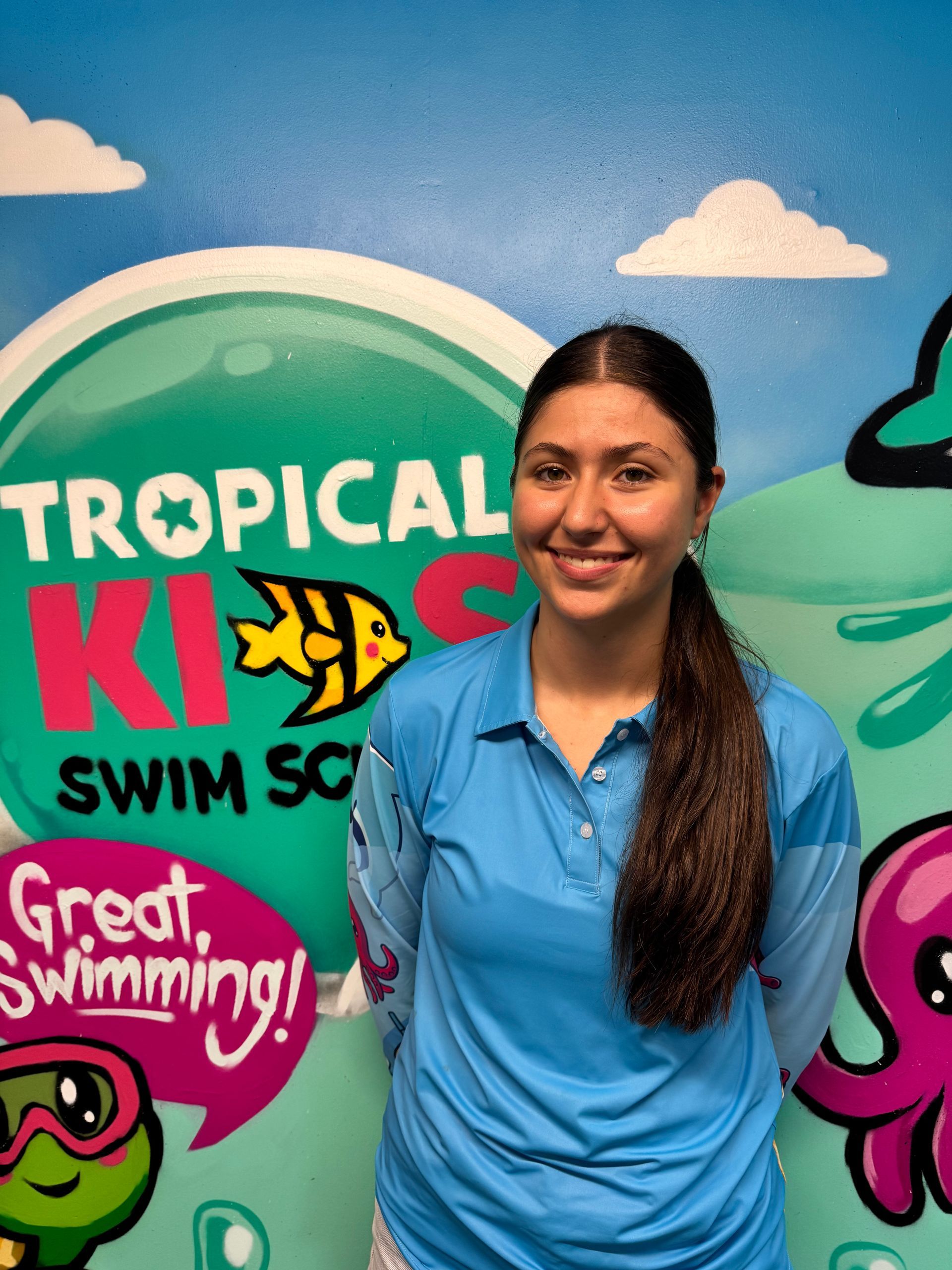 Person smiling in a pool, wearing a cap and blue shirt, standing in clear green water.
