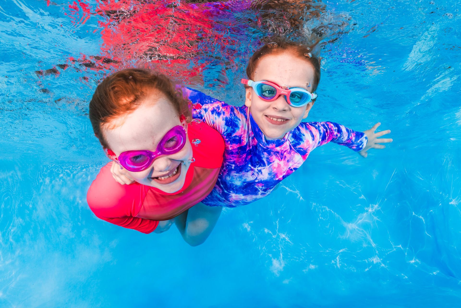 Two children in colorful swim goggles smiling in a bright blue pool
