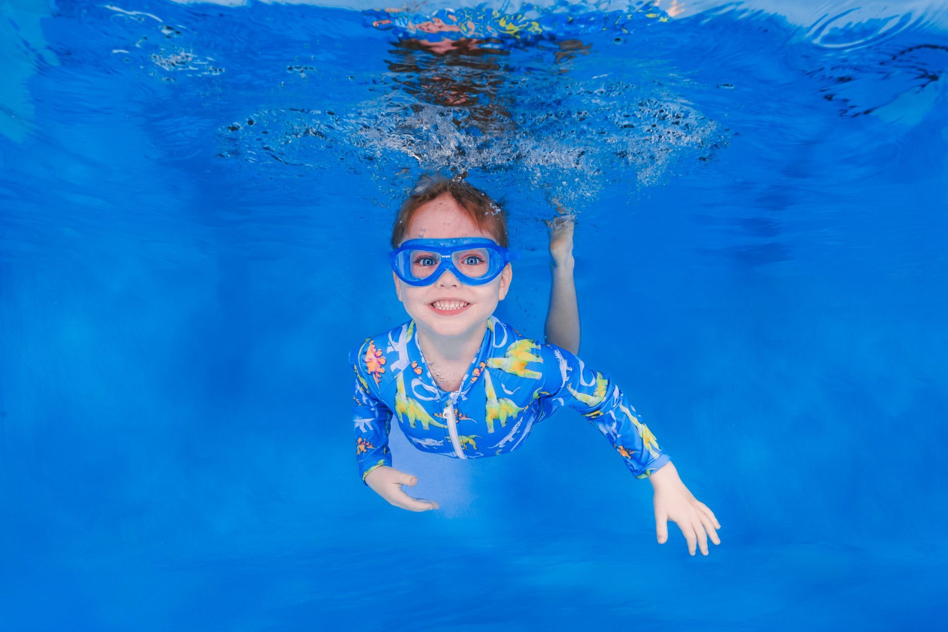 Child swimming underwater in a blue pool, wearing blue goggles and a blue floral rash guard.