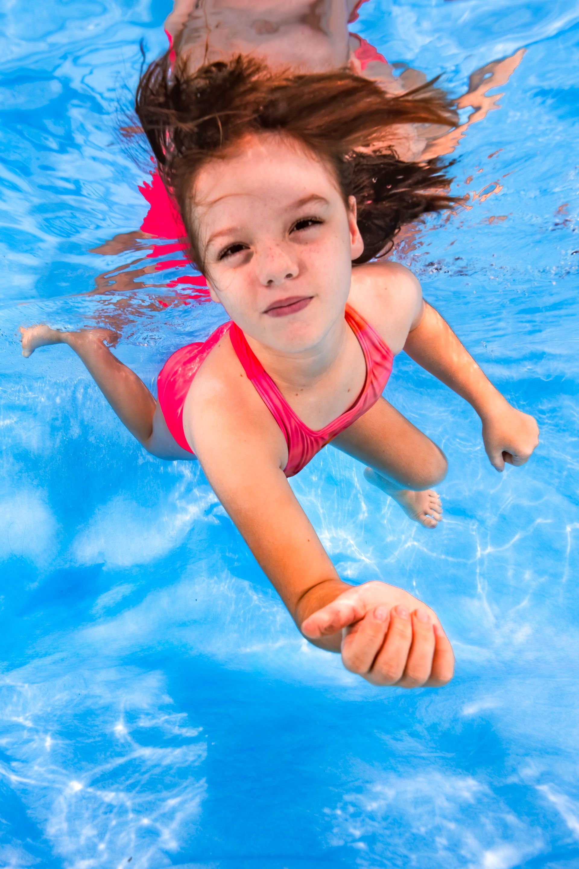 Child swimming underwater in a bright blue pool, wearing a pink swimsuit and reaching forward