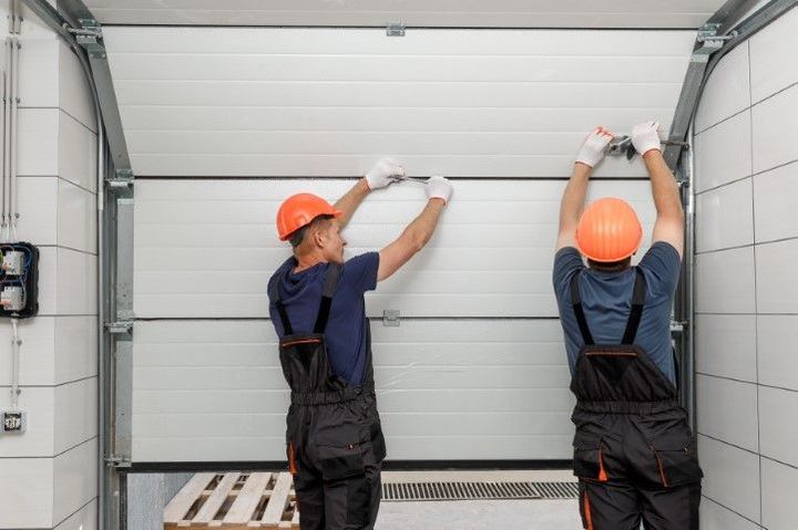 Two workers in orange hard hats lifting a garage door in a warehouse.