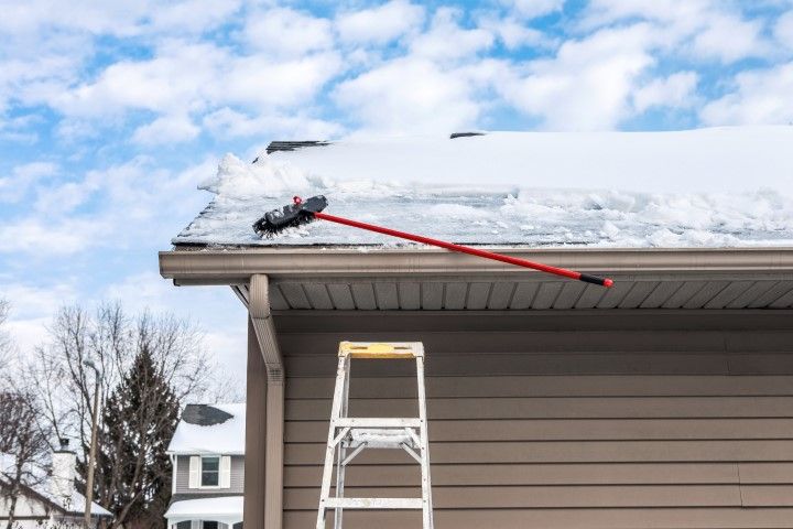 Person clearing snow from a roof with a red snow rake beside a ladder.