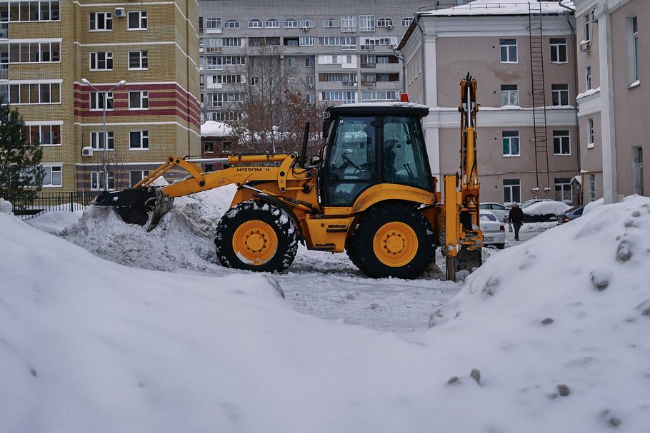 Yellow backhoe clearing snow on a city street between apartment buildings
