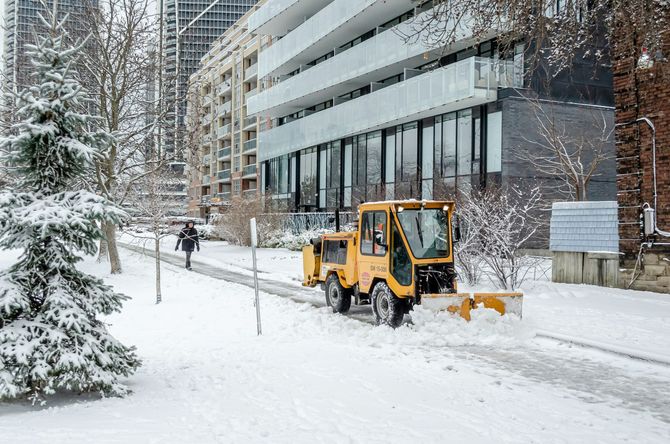 Yellow snowplow clearing a snowy sidewalk beside a modern building