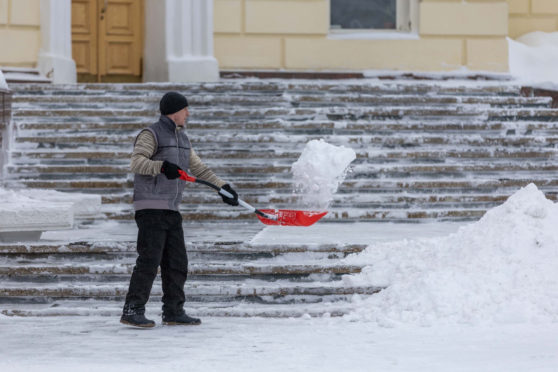 Person shoveling snow on steps outside a building, pushing a red snow shovel.