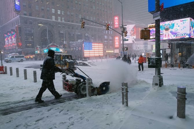 Person clearing heavy snow with a snowblower on a city street at dusk, with lit signs and traffic lights.