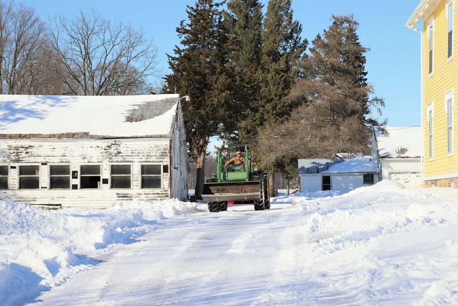 Snowy driveway with a green tractor parked between a garage and a yellow building