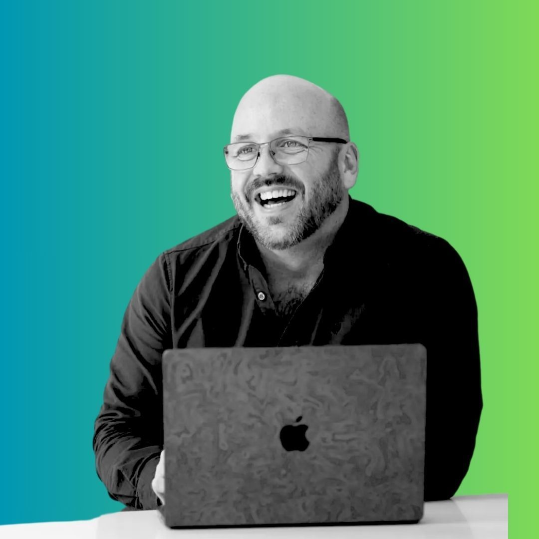 Smiling bald man in glasses behind a laptop on a teal-to-green background