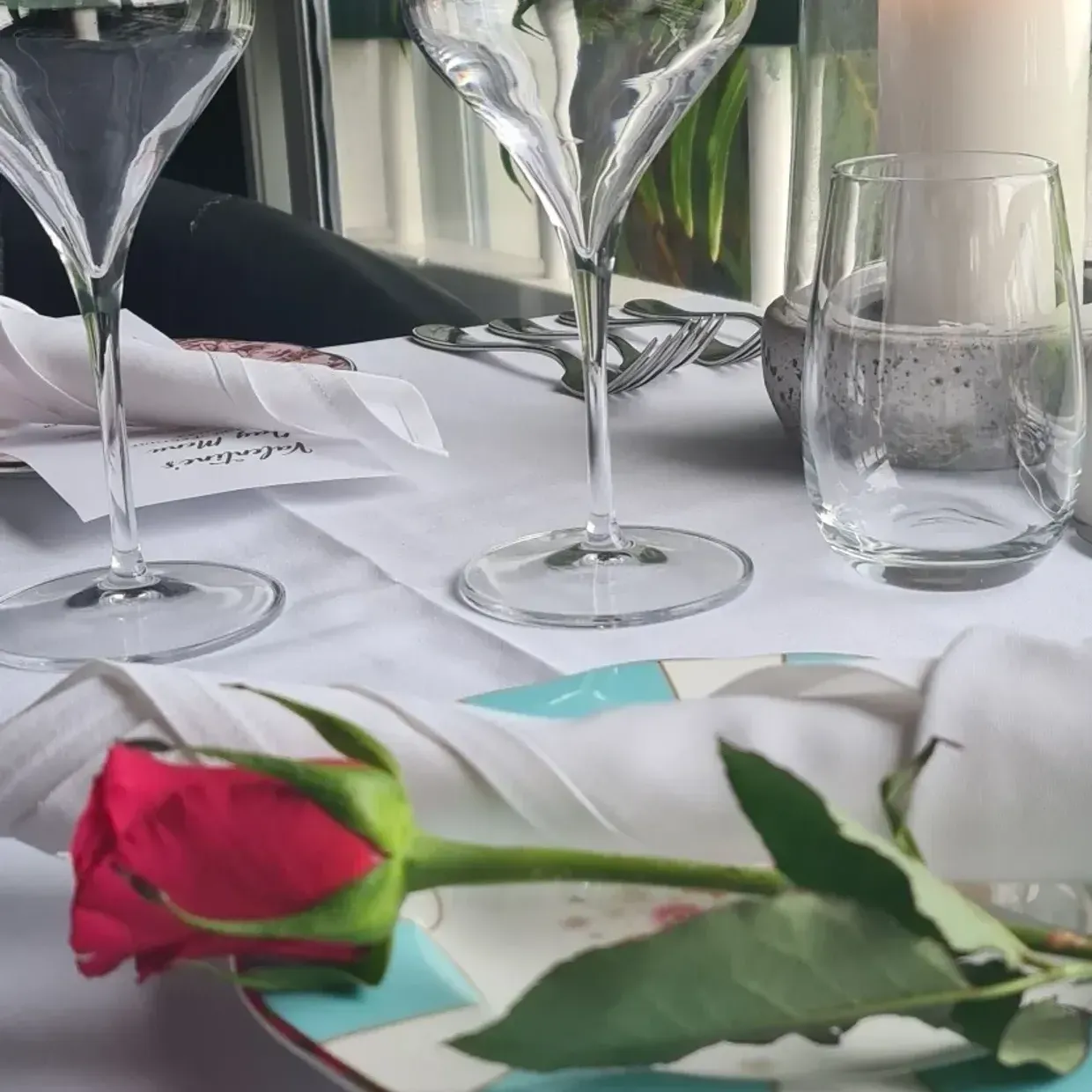A red rose sits on a plate next to wine glasses and a candle