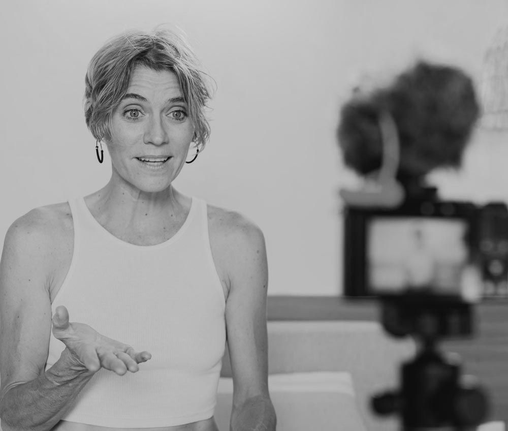 Woman in white top speaking to camera, with a camcorder blurred in the foreground.