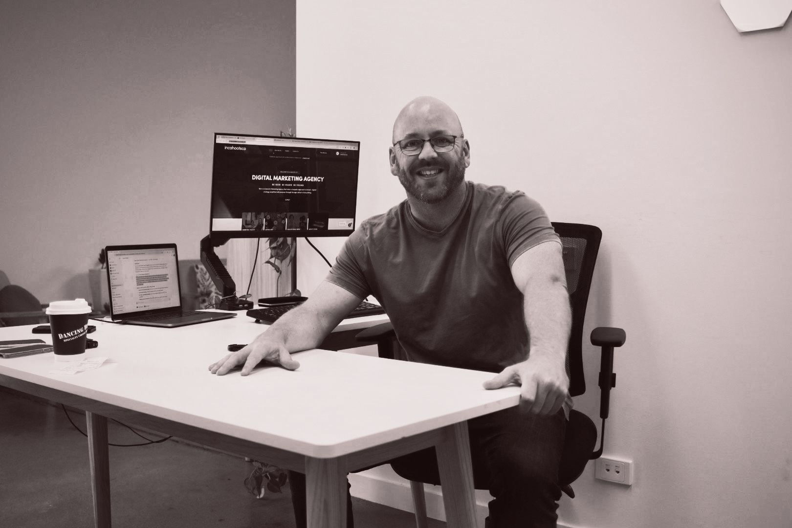 Smiling man seated at a desk in an office, with a computer monitor and paperwork nearby.