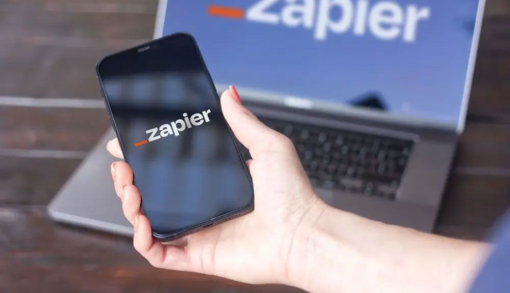 Hand holding a smartphone with Zapier logo in front of a laptop showing Zapier branding