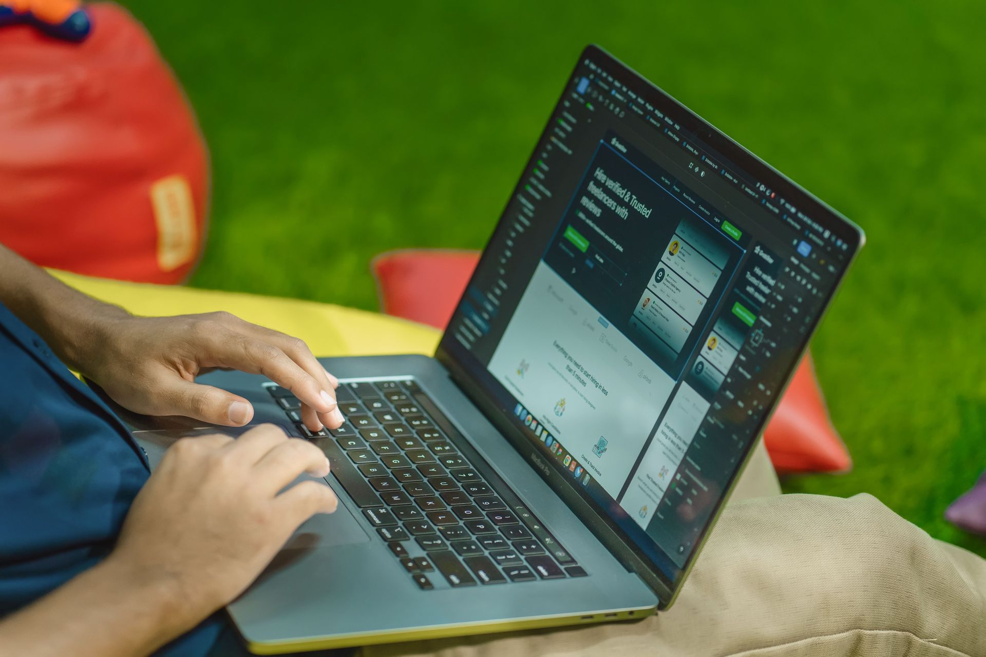 Person using a laptop outdoors on grass, with a red bag nearby and a screen showing software.