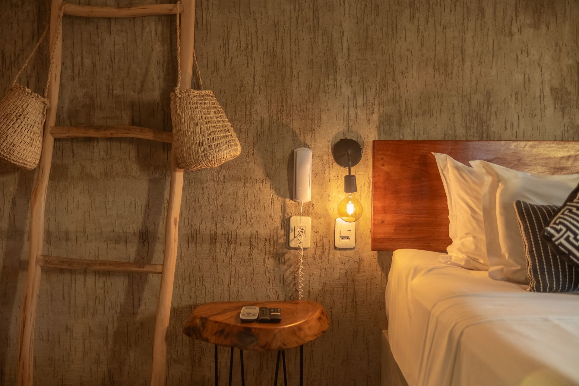 Um quarto aconchegante com uma parede texturizada, escada de madeira, uma pequena mesa lateral feita de tronco de árvore e um abajur de parede com luz quente perto da cama.