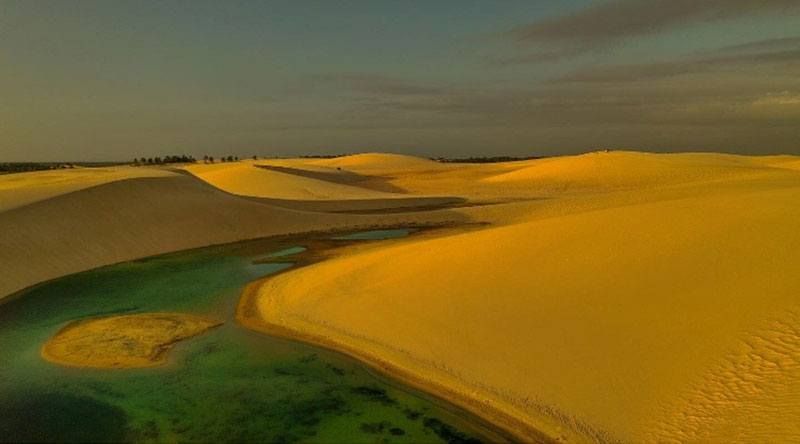 Dunas de areia dourada e brilhante circundam uma lagoa calma e turquesa sob um céu suave e crepuscular em Lençóis Maranhenses, Brasil.