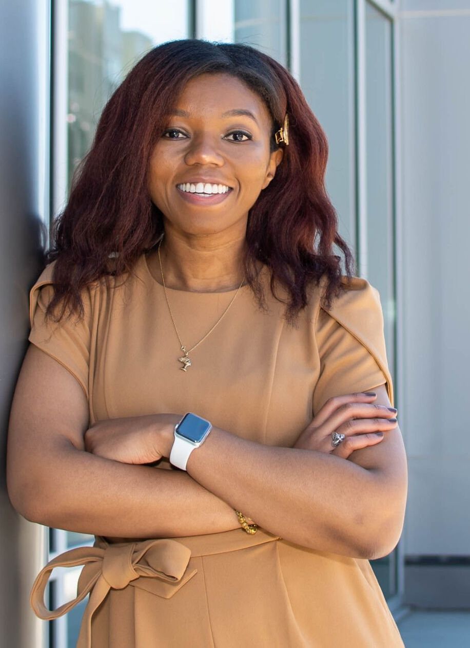 A woman in a brown dress is standing with her arms crossed and smiling.