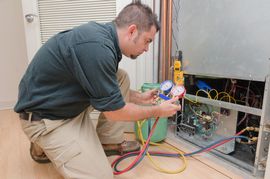 A technician kneels while using a manifold gauge set to check a residential HVAC unit inside a utility room.