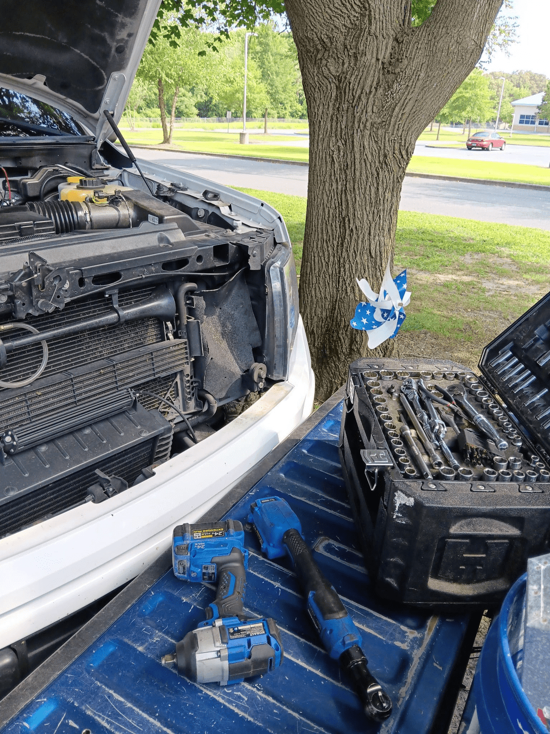 Truck with open hood, tools in bed, being repaired outdoors near a tree.
