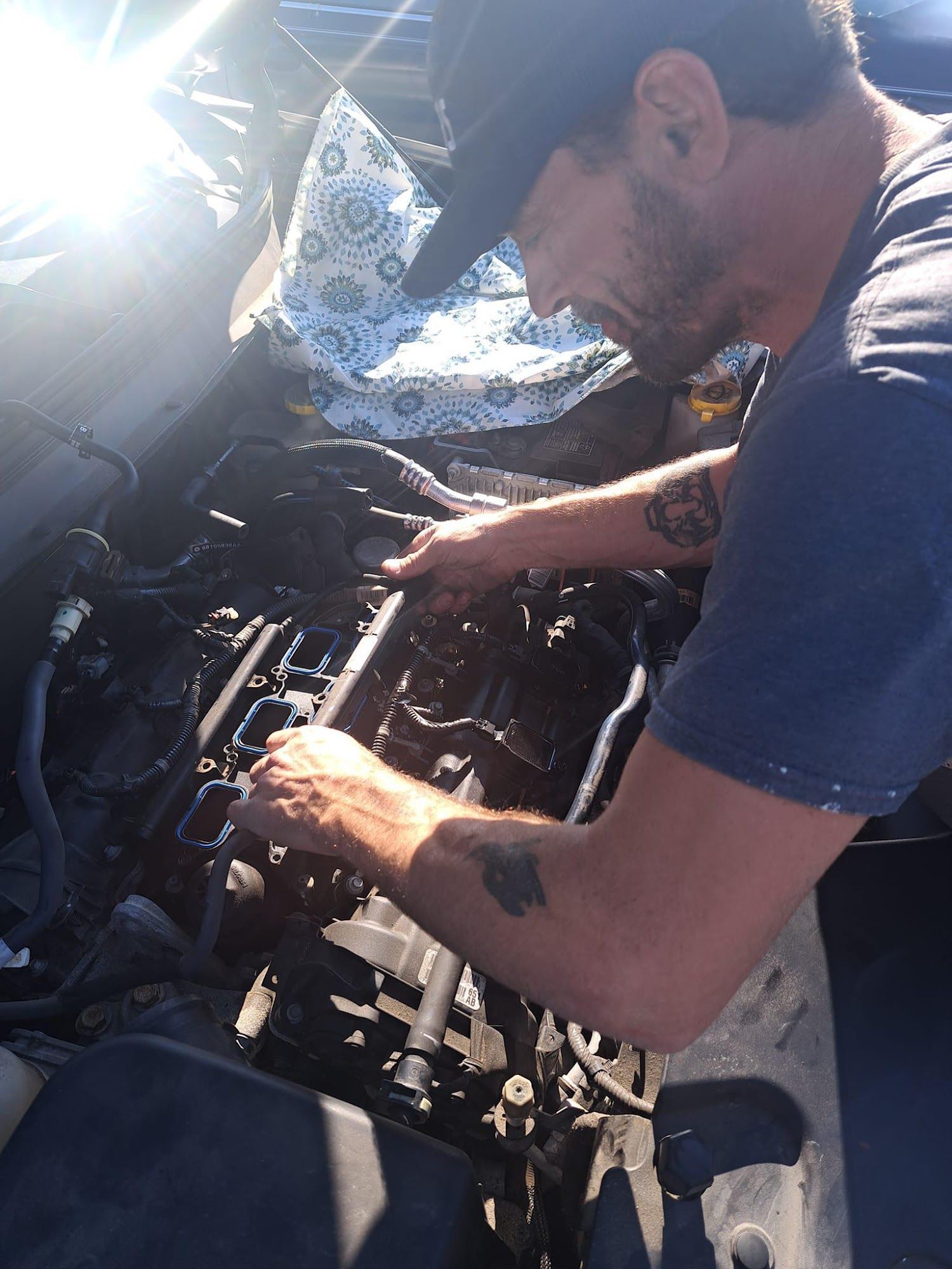 Man working on a car engine, wearing a hat. He is using tools. Blue shirt, sunny setting.