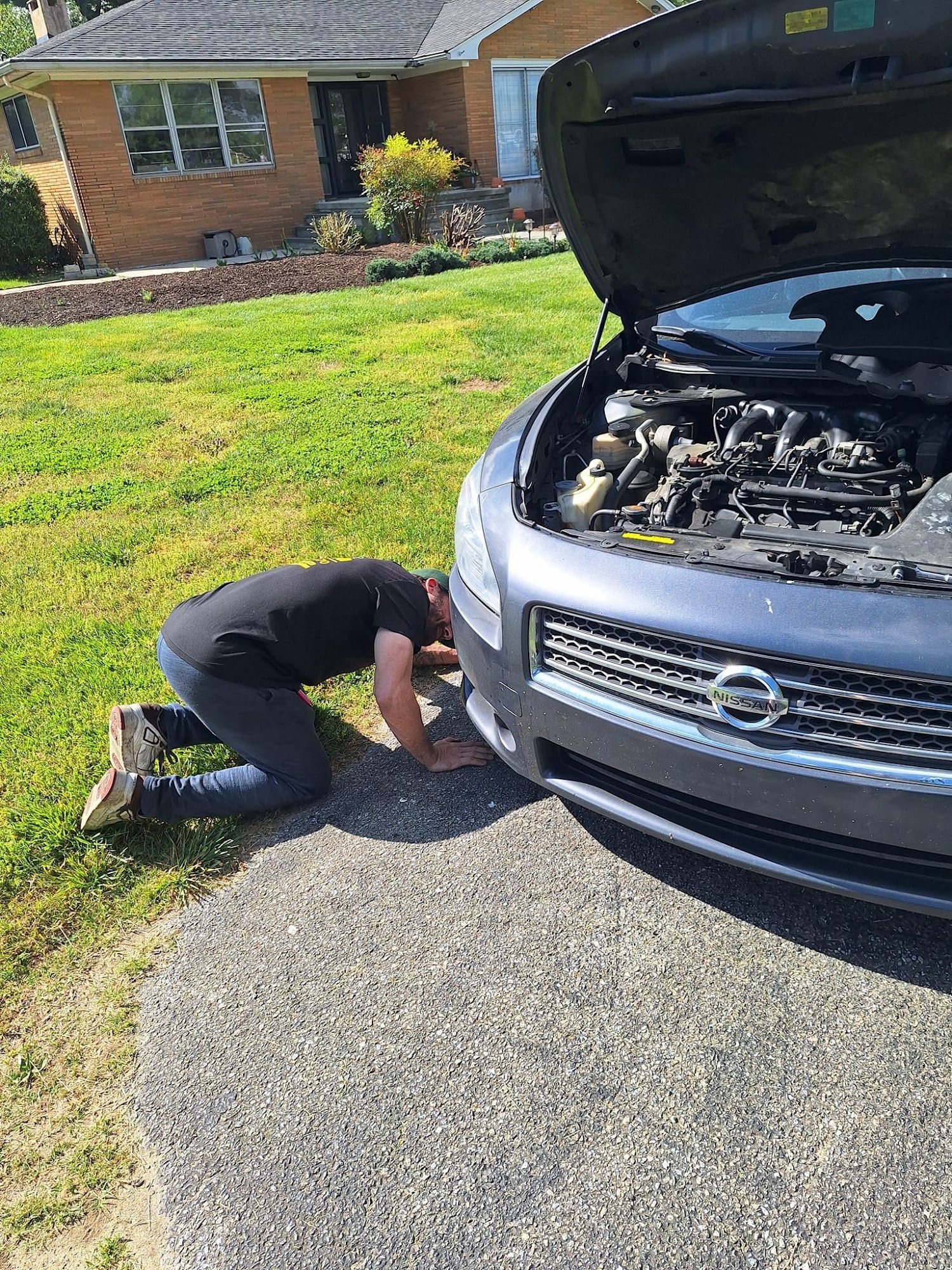 Person kneeling in front of a blue car with hood open, working on the engine, in a yard.