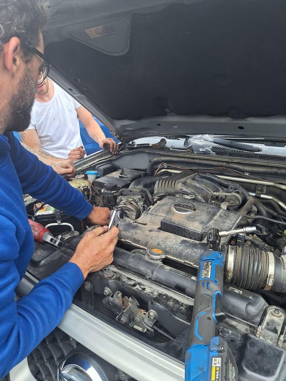 Two people work on a car engine with tools, outdoors. One wears blue, the other white.