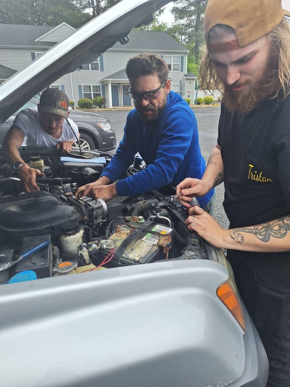 Three people working on a car engine outdoors. The hood is open. One is wearing a blue jacket, others t-shirts.