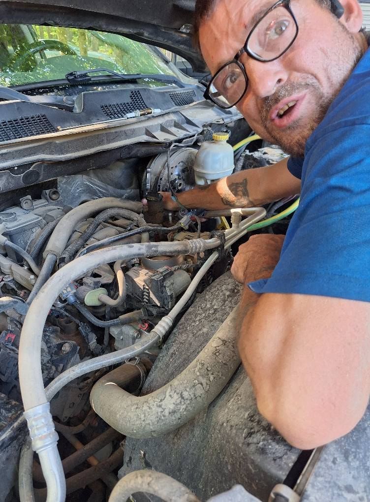 Man working on a car engine, looking surprised, wearing glasses and blue shirt, outdoors.