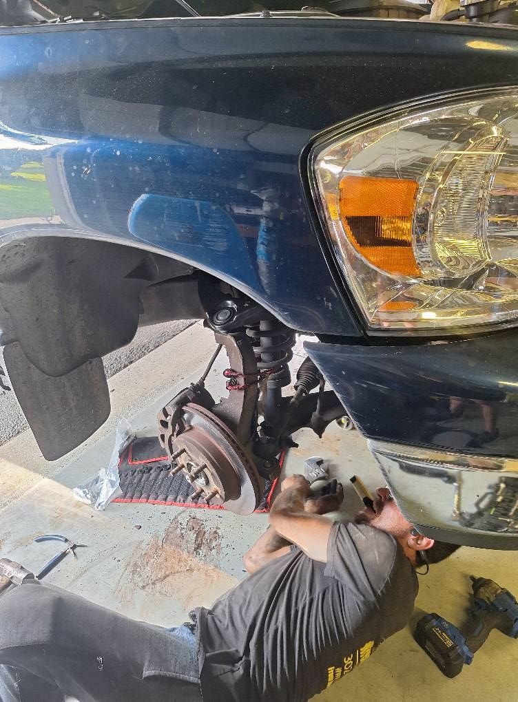 Mechanic working under a blue truck, removing a tire. Visible front suspension parts and tools.