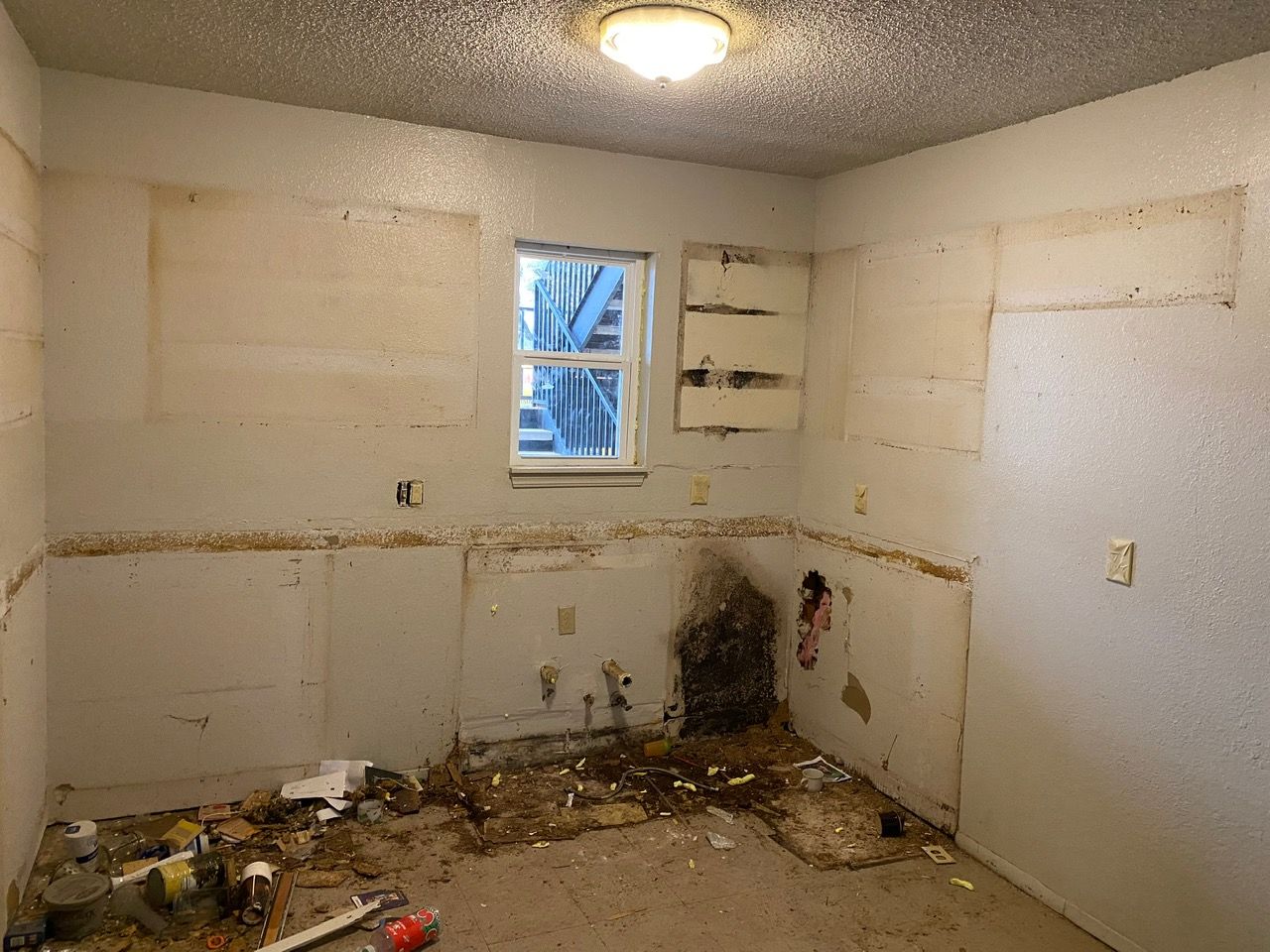 Interior room gutted, walls stripped, debris on floor; window with scaffolding visible.