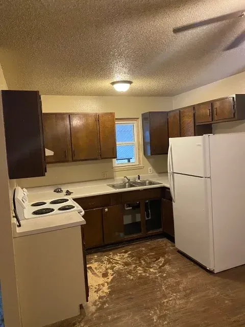 A kitchen with brown cabinets, white appliances, and damaged flooring.
