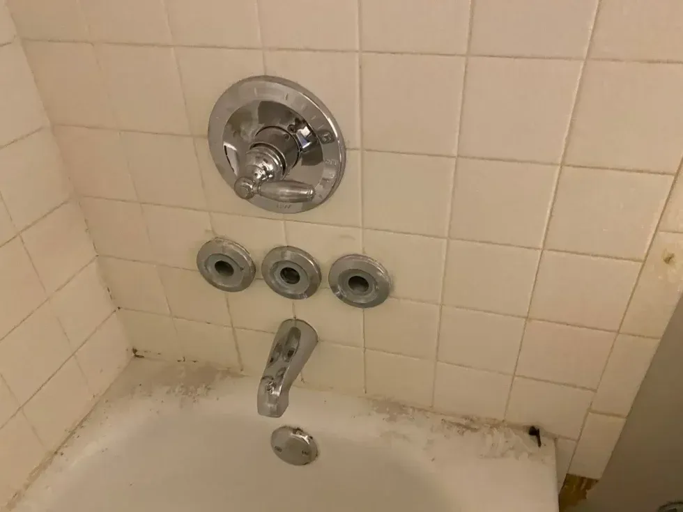 A bathtub faucet and shower controls on a tiled wall, showing signs of mildew and wear.