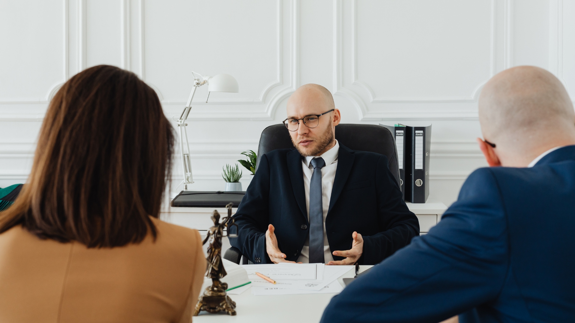 A lawyer sitting at a desk and gesturing while speaking to two clients in a white office.