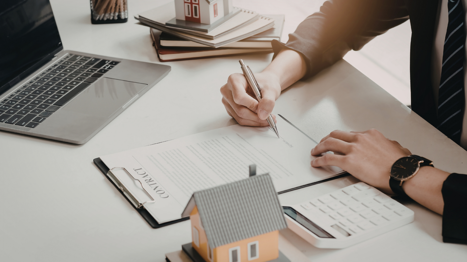 A person in a suit signs a real estate contract at a desk with a laptop, calculator, and a small model house.