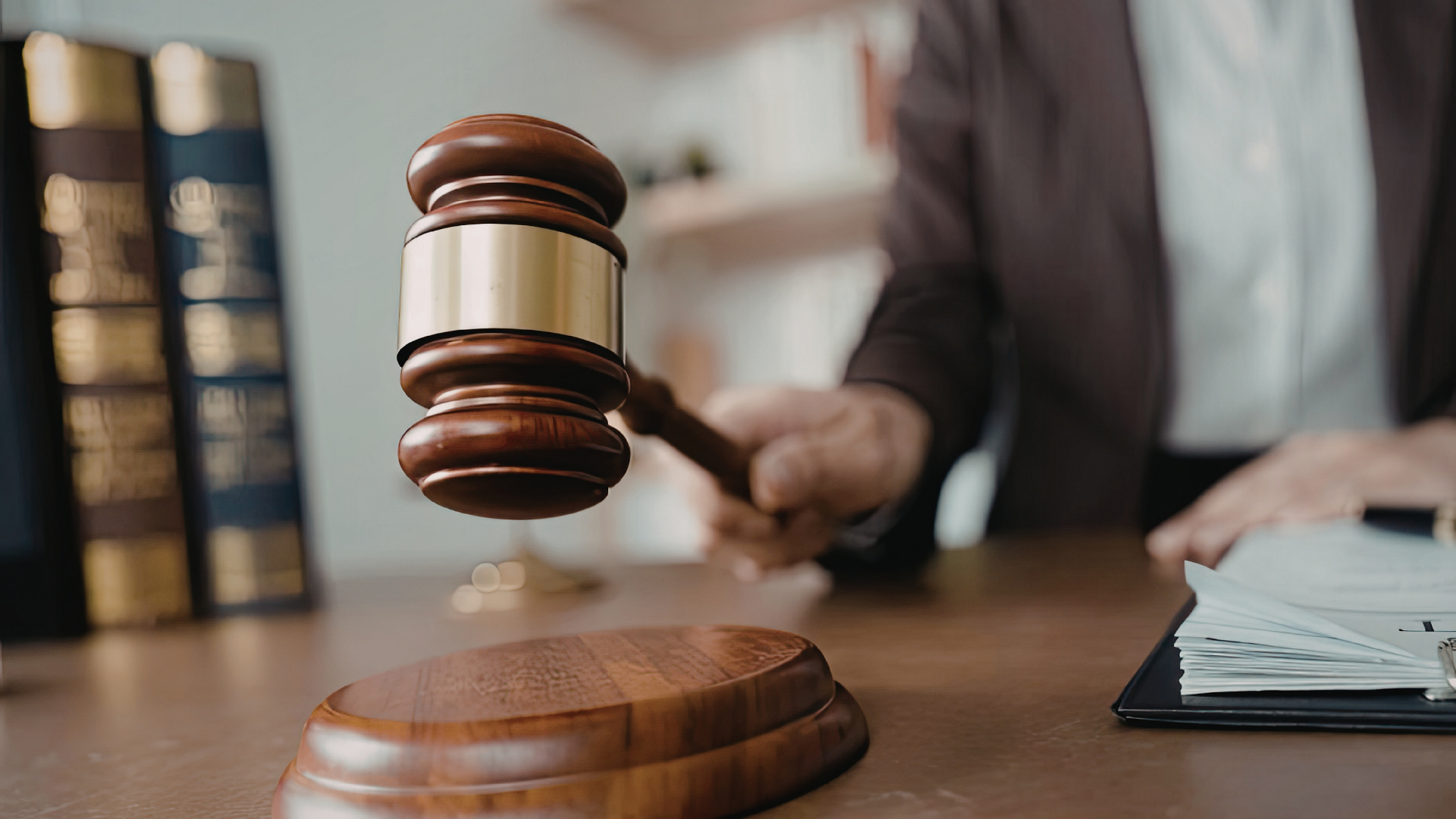 A wooden judge's gavel held above a sounding block in a courtroom with law books and papers on a desk.