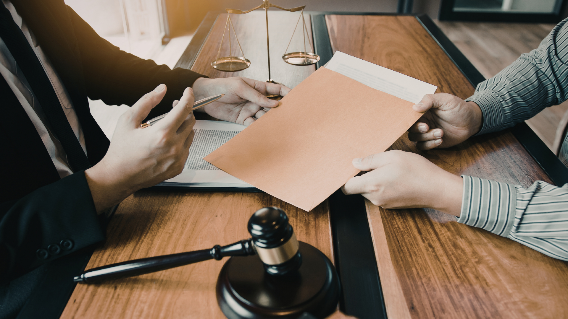 A person in a suit hands a document to another person across a wooden table with a judge’s gavel