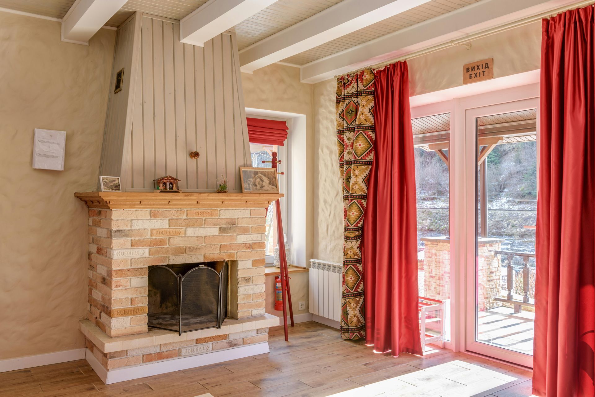 Bright living room with brick fireplace, tall white curtains, and red drapes beside glass doors.