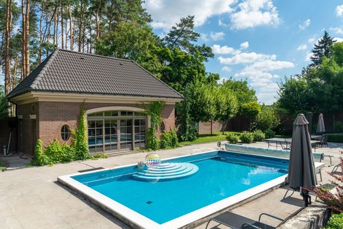 Backyard pool with blue water beside a brick patio and small building, surrounded by trees and umbrellas