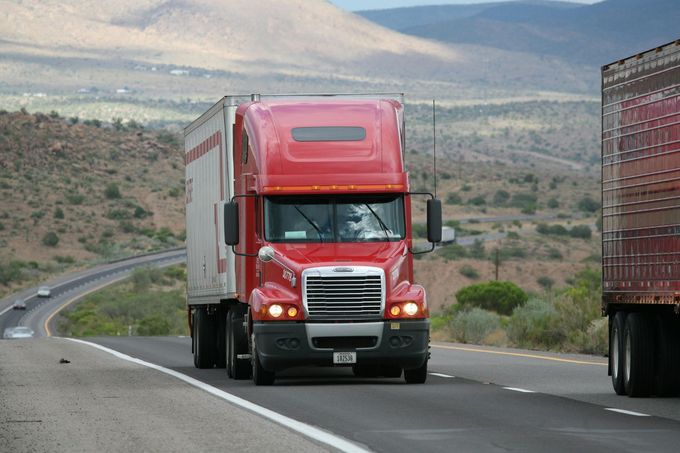 Red semi-truck driving on a highway, another truck to the right, mountains in the background.