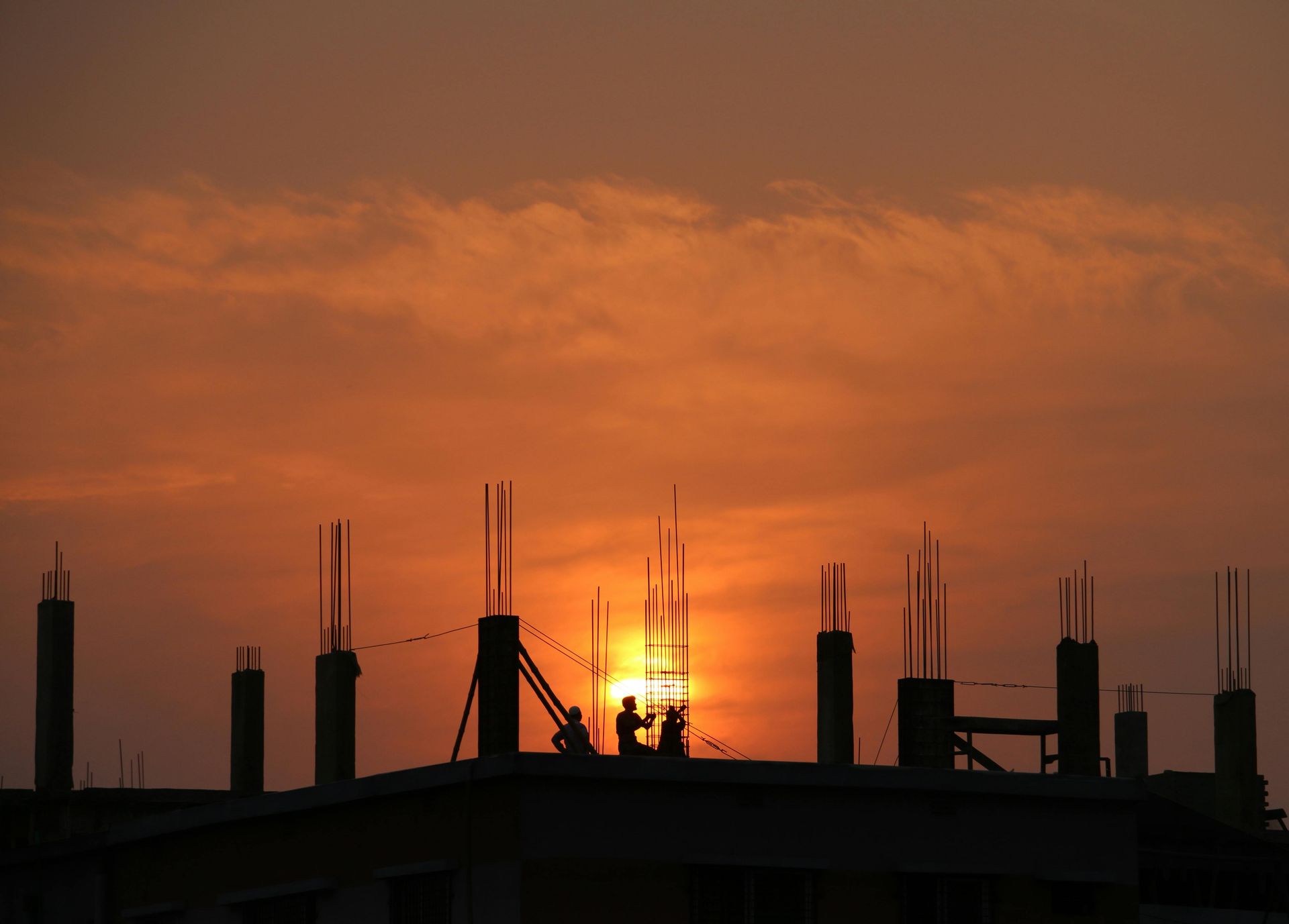 Construction site silhouette against orange sunset; workers stand atop structure.