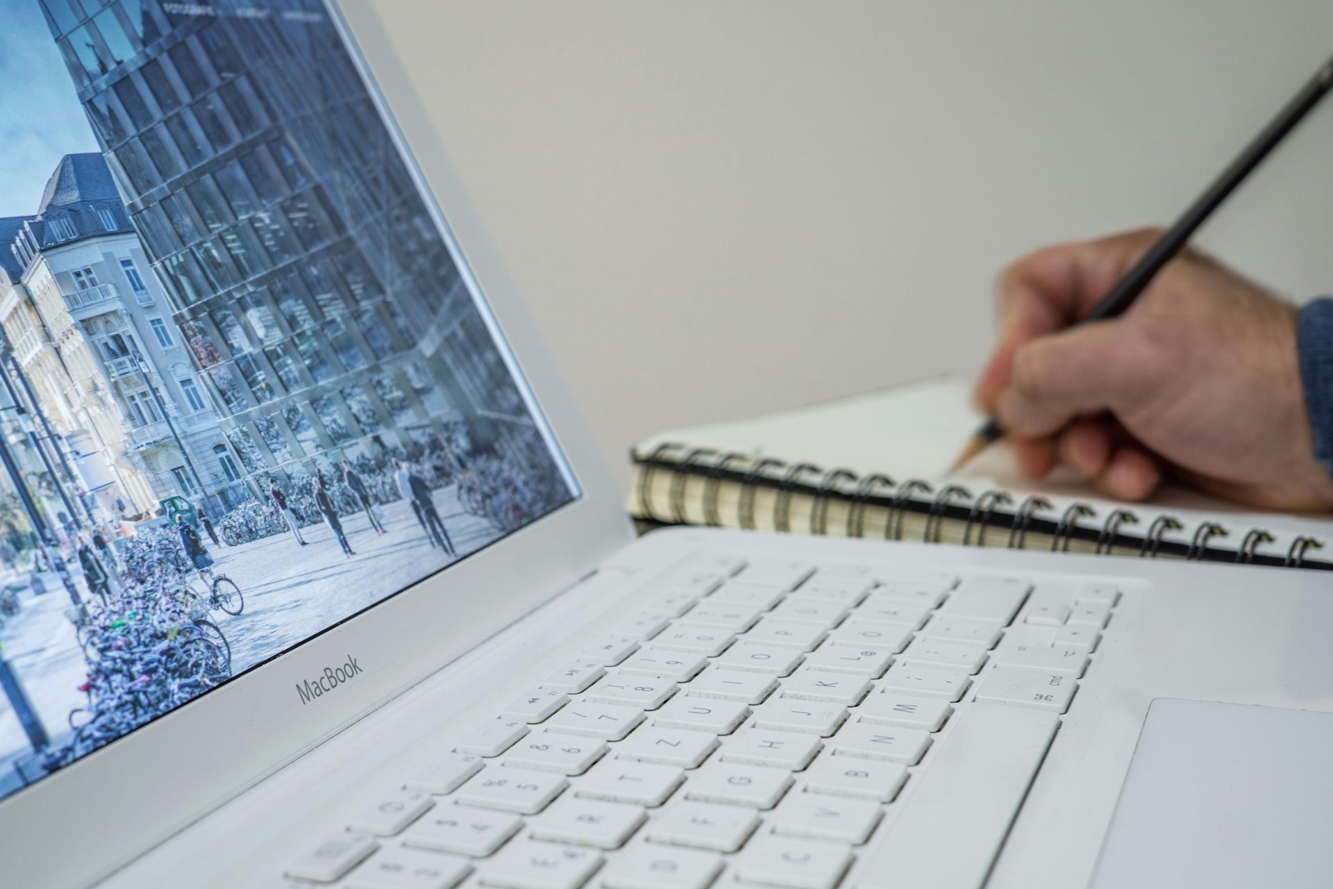 White laptop displays city scene; person writes in notebook with pencil.