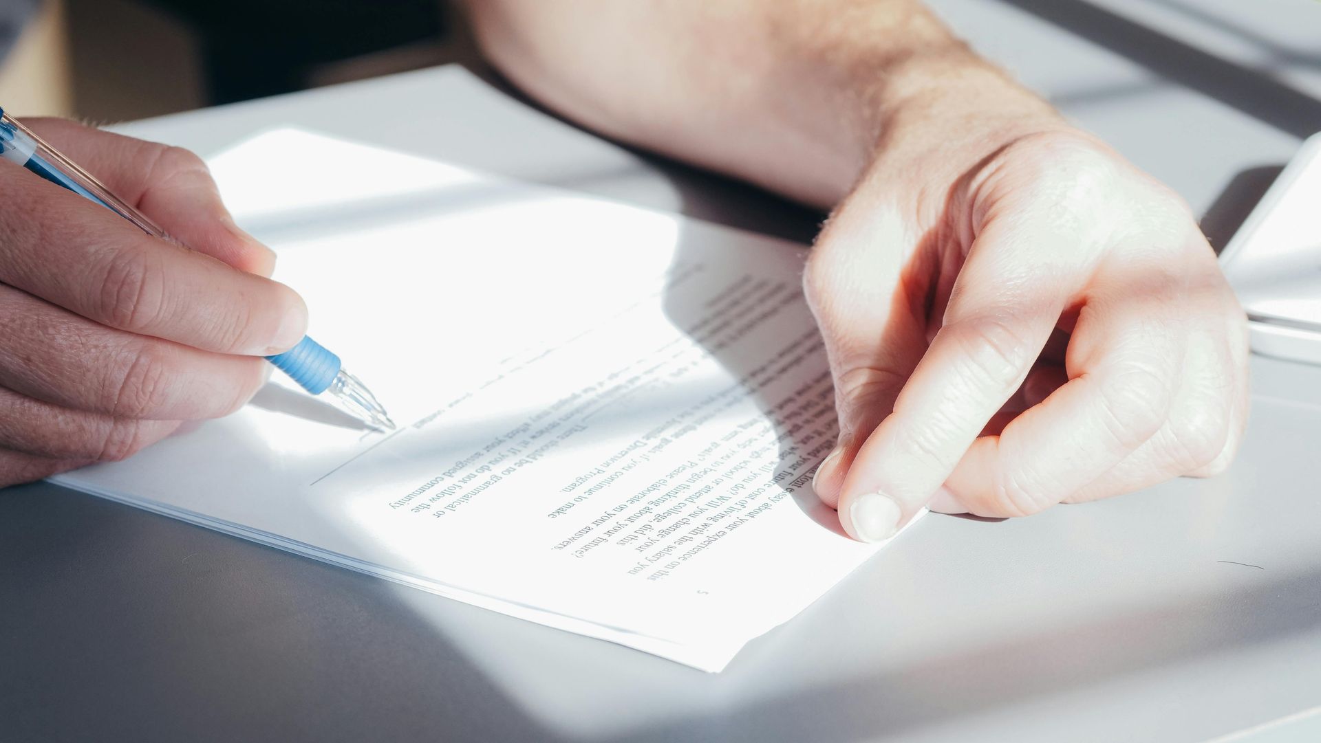 Person's hands writing with a blue pen on a white document on a gray surface.