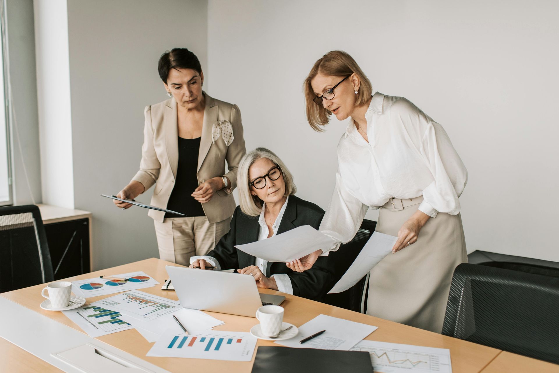 Three women review documents and a laptop at a table in an office setting.