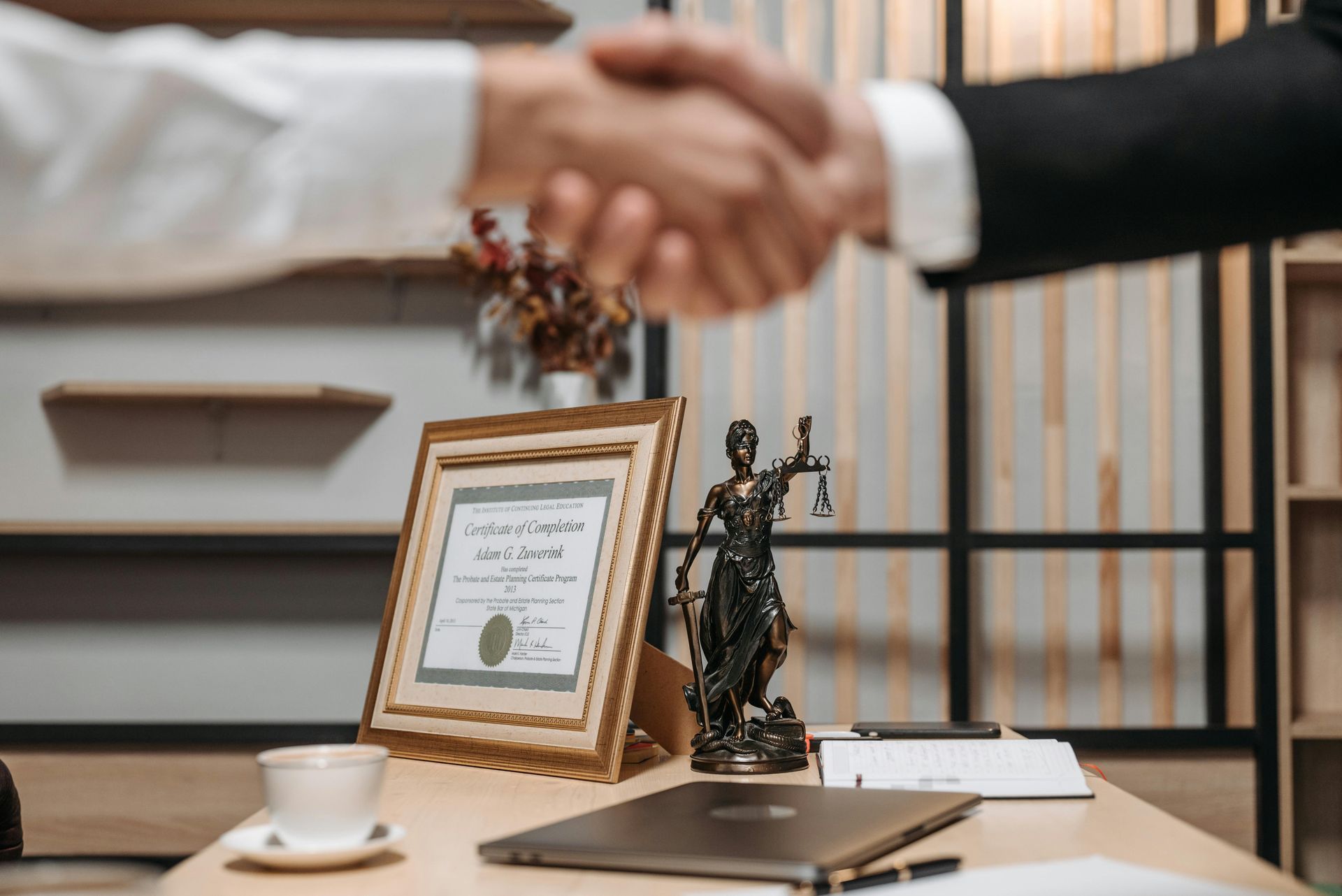 Two people shake hands near a desk with a law office setting, statue of justice and a certificate.